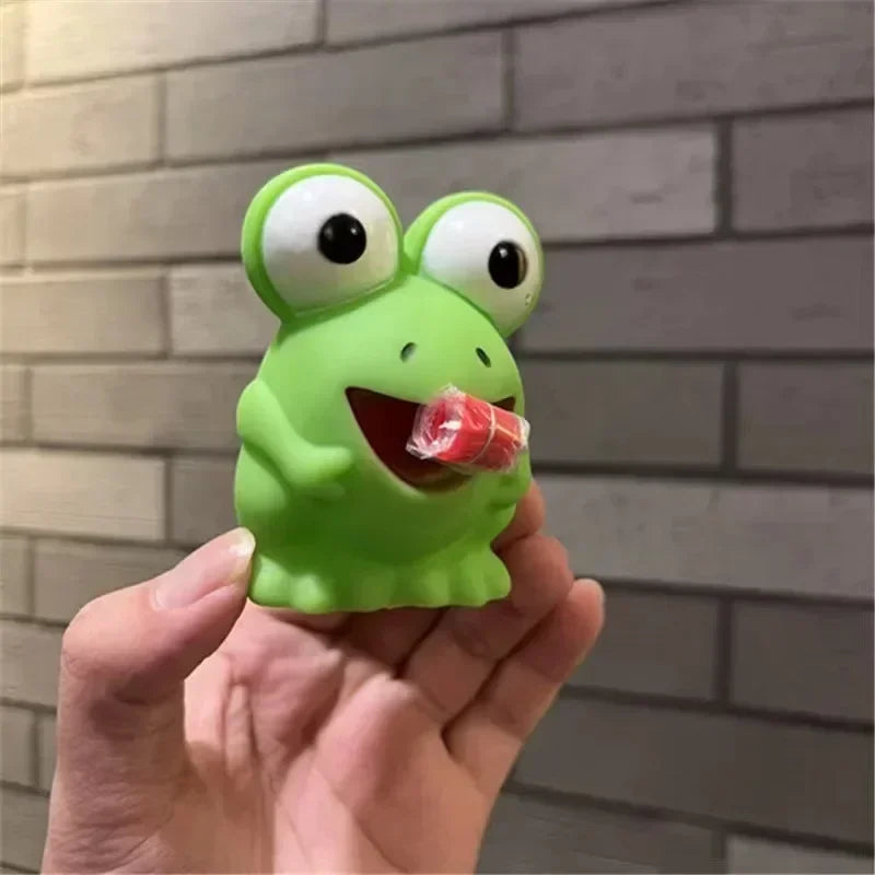 Green frog-shaped object with a candy in its mouth held by a hand against a brick wall.